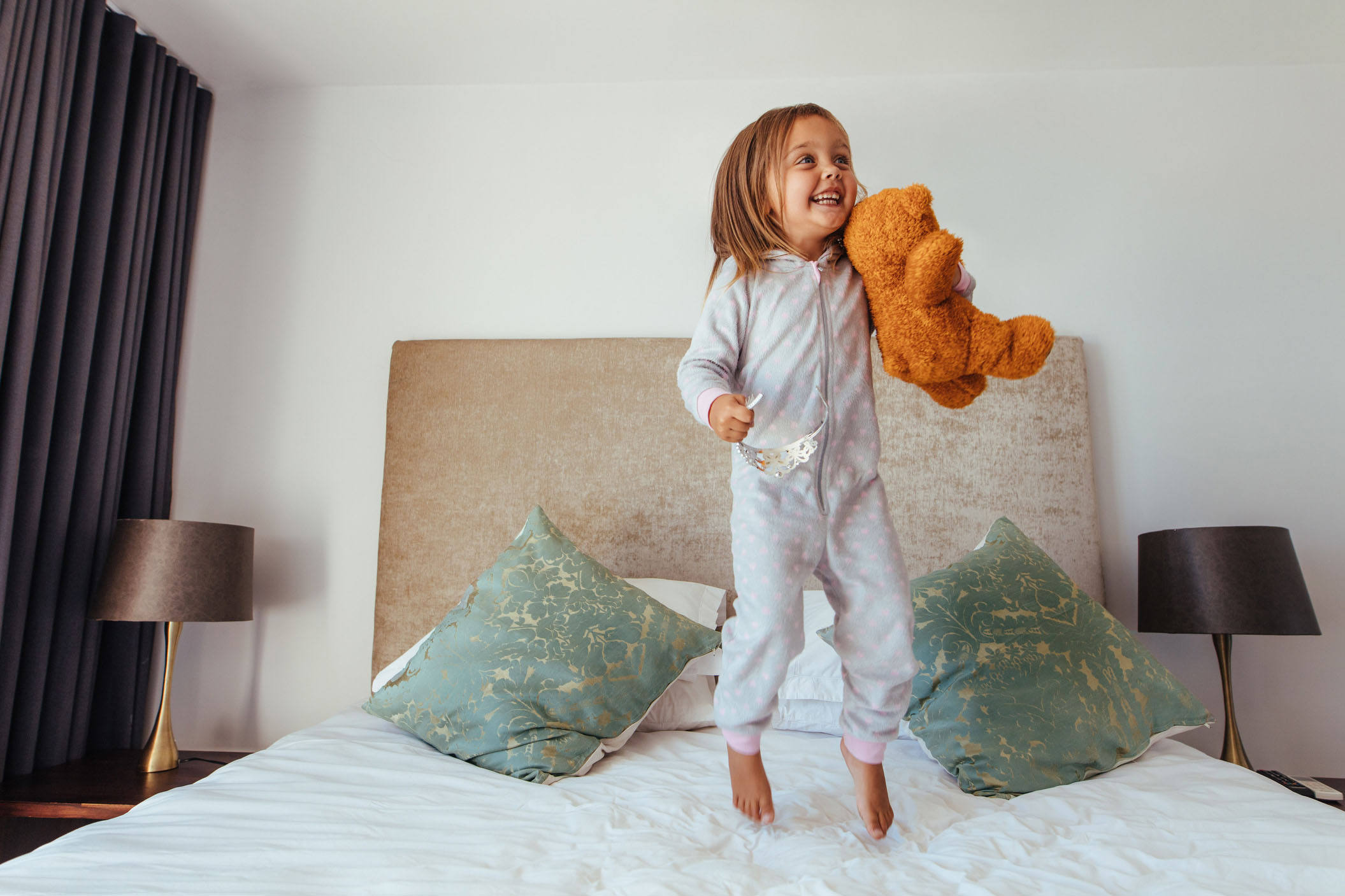 Smiling young girl jumping on a freshly cleaned bed with teddy bear — non-toxic mattress cleaning safe for kids and pets in Orange County.